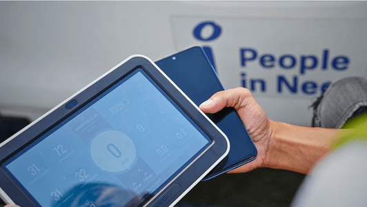 A person holds a tablet and a mobile near a sign that reads "People in Need.