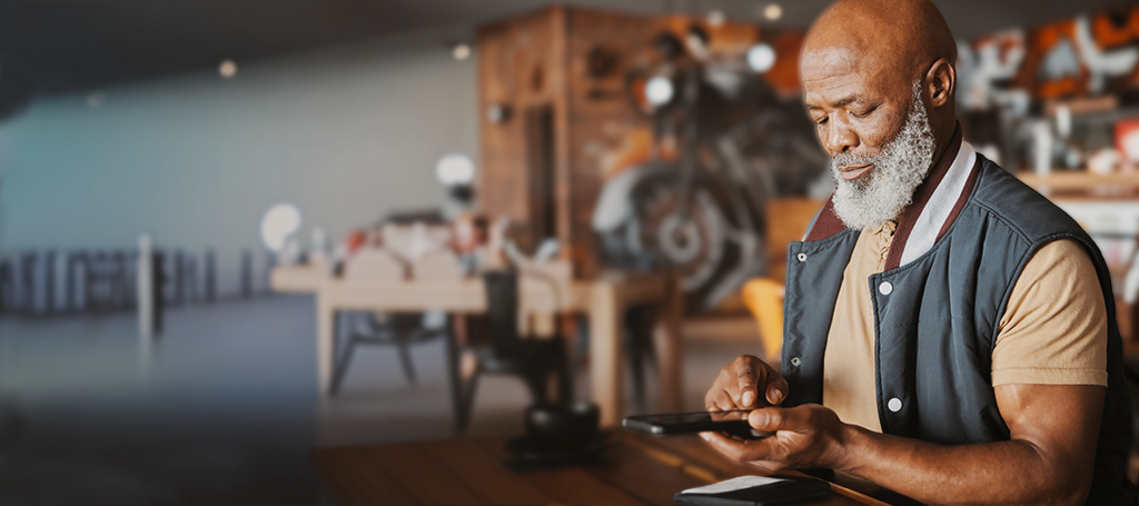 Older man using a mobile phone at a wooden table in a modern, cosy café with blurred background.