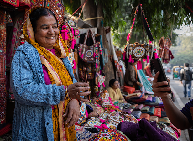 A woman in colourful clothing smiles for a photo at a vibrant outdoor market with embroidered bags.