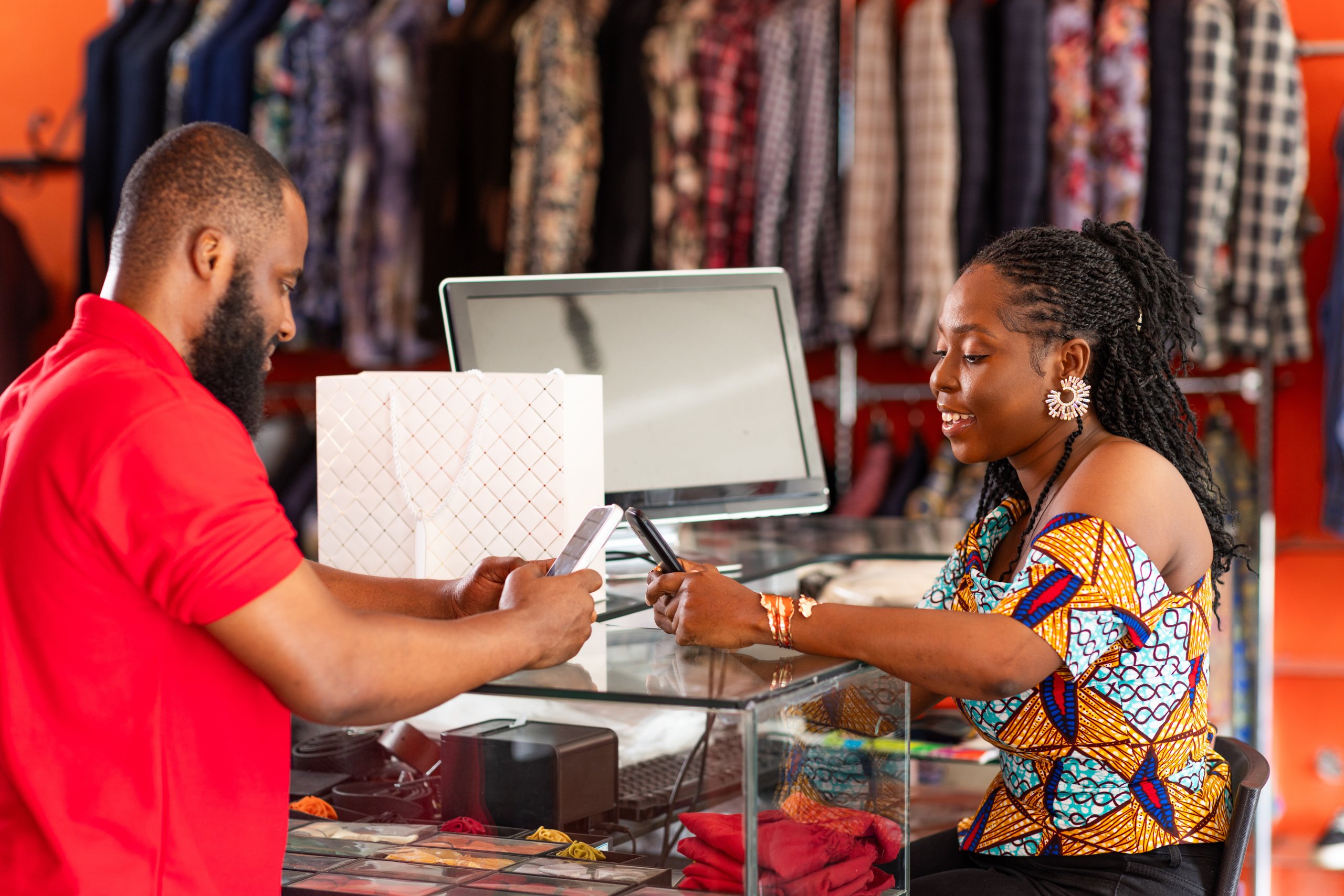 A woman at a boutique counter pays a cashier using her mobile, with clothes hanging in the background.
