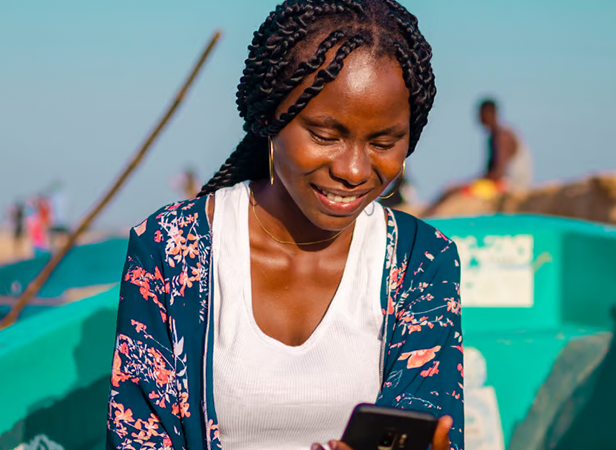 An anticipatory smile spreads across the woman's face as she looks at her mobile, sitting outdoors near a turquoise boat with her plaits gently swaying.