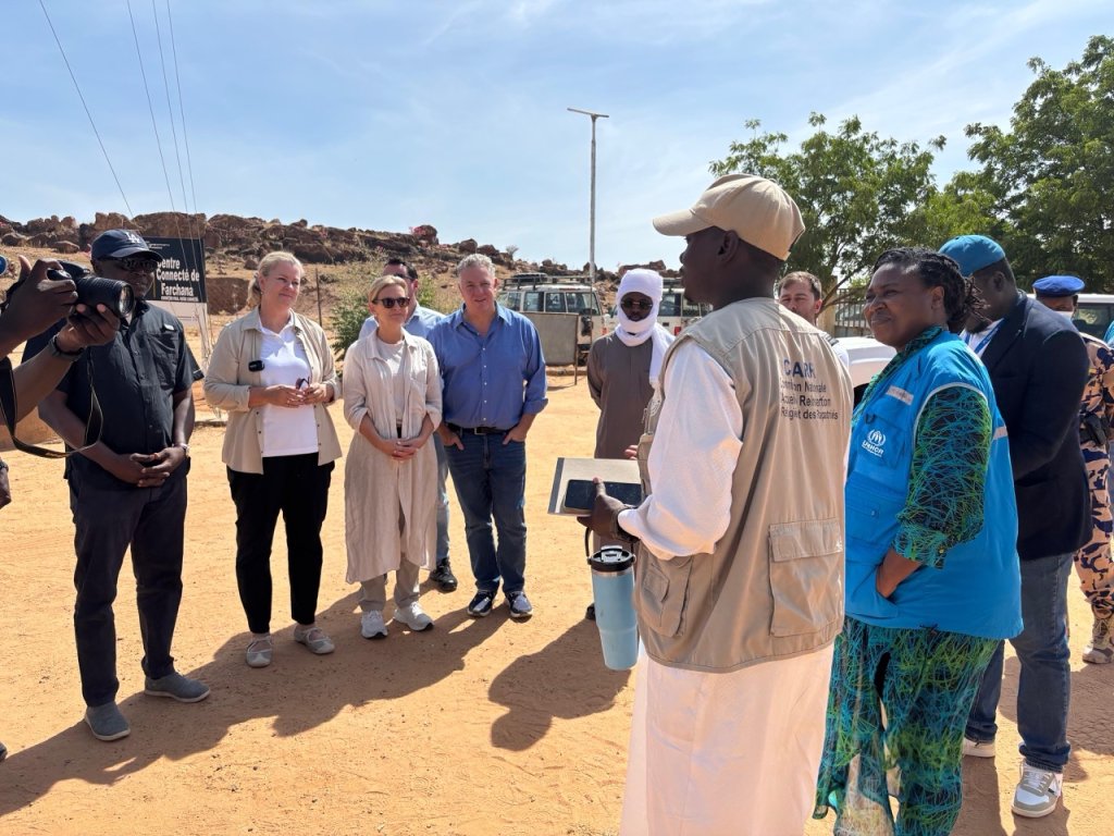 A group of aid workers and officials stand outdoors, listening to a man speaking in a sunlit area.
