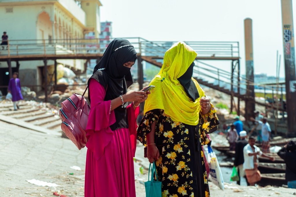 Two women in hijabs stand outdoors; one in pink checks her mobile, the other in yellow carries a shopping bag.