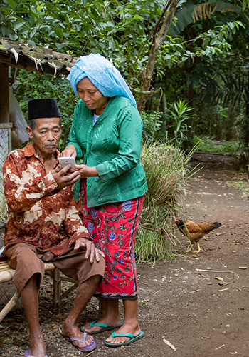 An elderly man and a woman look at a mobile phone together outdoors, with greenery and a hen in the background.