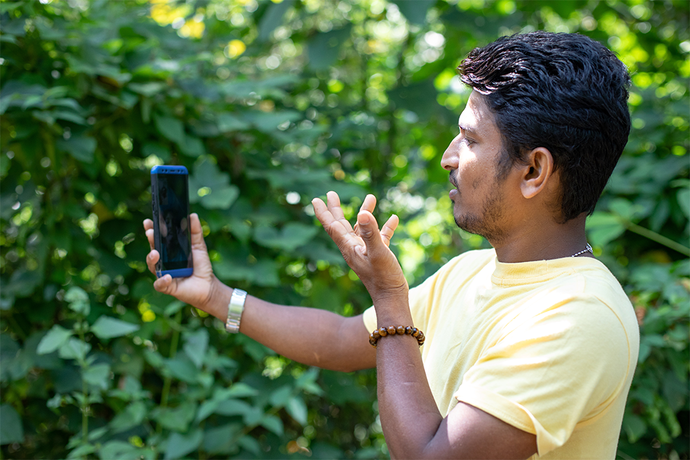 Man in yellow shirt video calling on mobile outdoors, gesturing with one hand, green foliage in background.