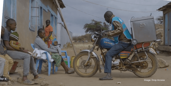An anticipatory man on a motorbike talks to a seated family outside a house in a rural area.