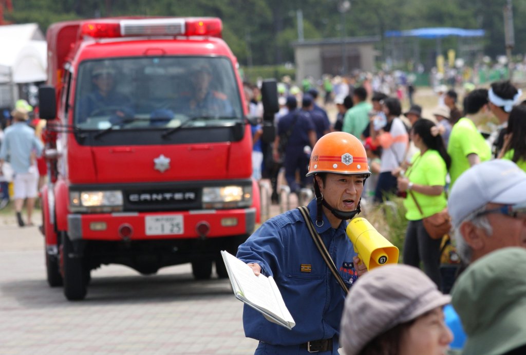 A firefighter with a helmet directs people near a fire engine and a crowd at an outdoor event.