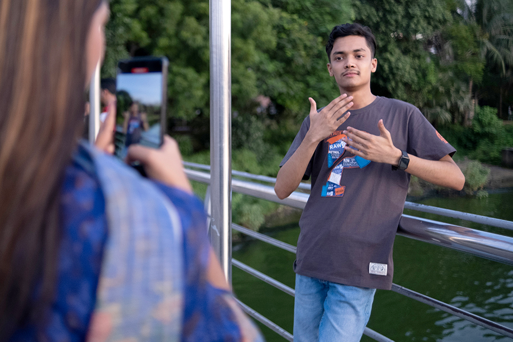 A young man poses on a bridge as someone takes his photo with a mobile phone near green trees and water.
