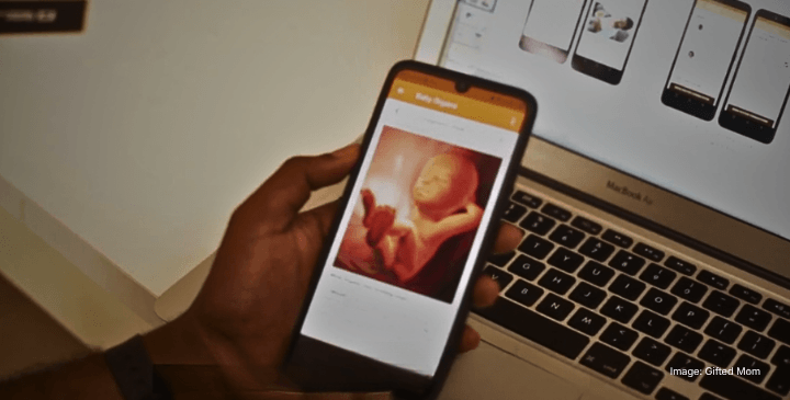 A hand holds a mobile displaying an anticipatory foetus image, with a laptop showing app designs in the background.