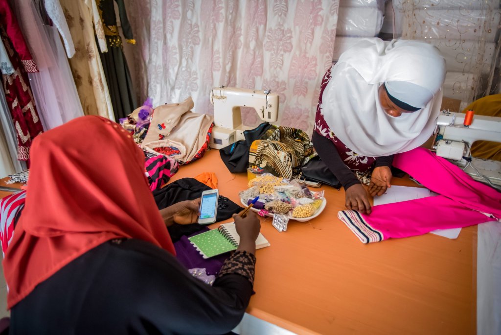 Two women sew fabric at a table with sewing machines, one using a phone and notebook, the other pinning fabric.