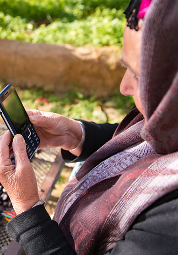 Woman in a headscarf holding and looking at a mobile phone outdoors in a garden setting.