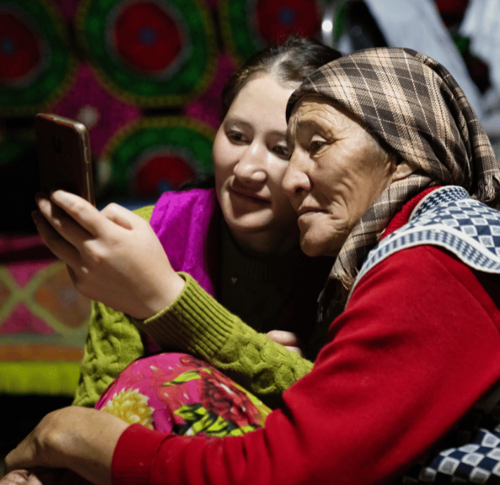A young woman and an older woman sit close together, smiling at a phone for a selfie, capturing the moment with the help of EmergingTech.