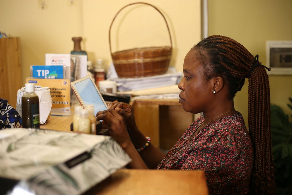 Woman with plaited hair uses a tablet whilst seated at a desk with various bottles and a basket in the background.