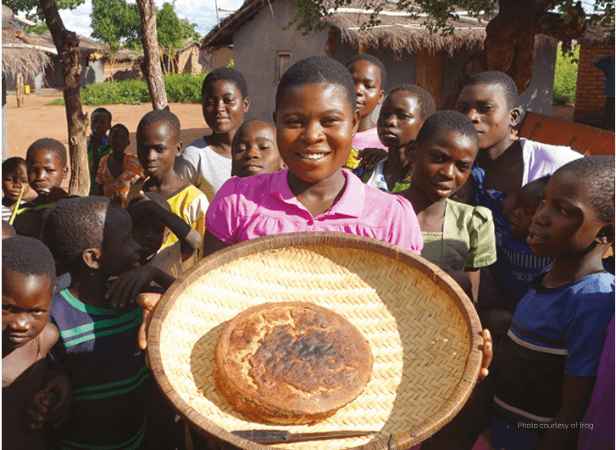 Smiling girl holding a round loaf on a woven tray, surrounded by children in an anticipatory mood in a rural village setting.