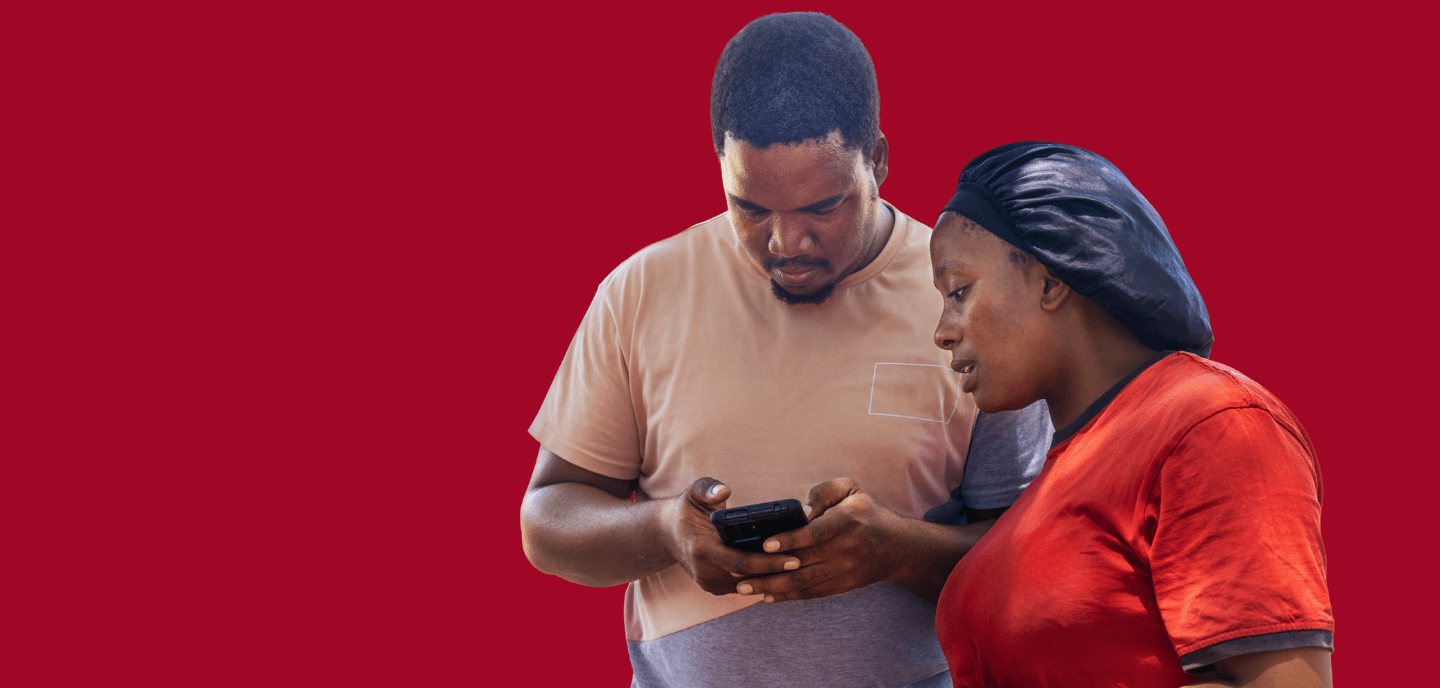 A man and woman look together at a mobile phone, symbolising digital humanitarian assistance, against a solid red background.