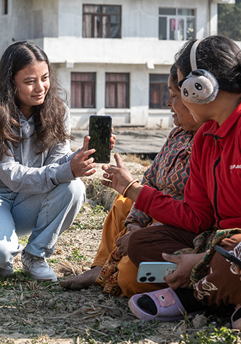 A young woman shows a mobile phone to two seated women outdoors; one woman wears headphones.