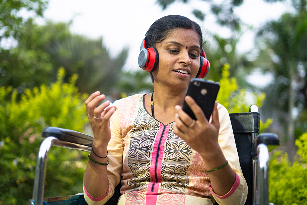 Woman in a wheelchair wearing headphones, smiling and looking at her mobile outdoors.