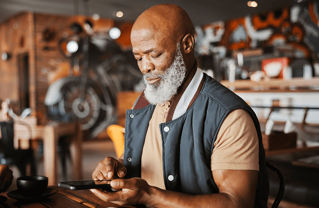 Older man with a grey beard using a mobile phone in a modern, art-filled café, exploring GSMA Mobile for Development initiatives.