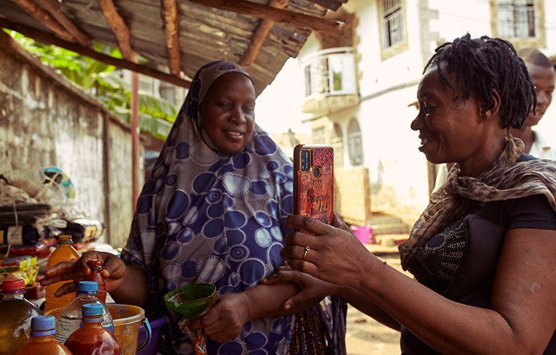 Two women at an outdoor market; one smiles whilst the other takes a photo of her with a mobile phone.