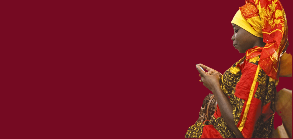 Anticipatory woman in colourful traditional attire sits and looks at her mobile against a plain maroon background.