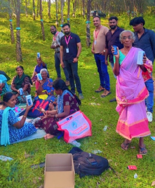 Elderly women in colourful sarees receive mobile phones outdoors, smiling with a group of people in a grassy area, celebrating handset affordability in India.
