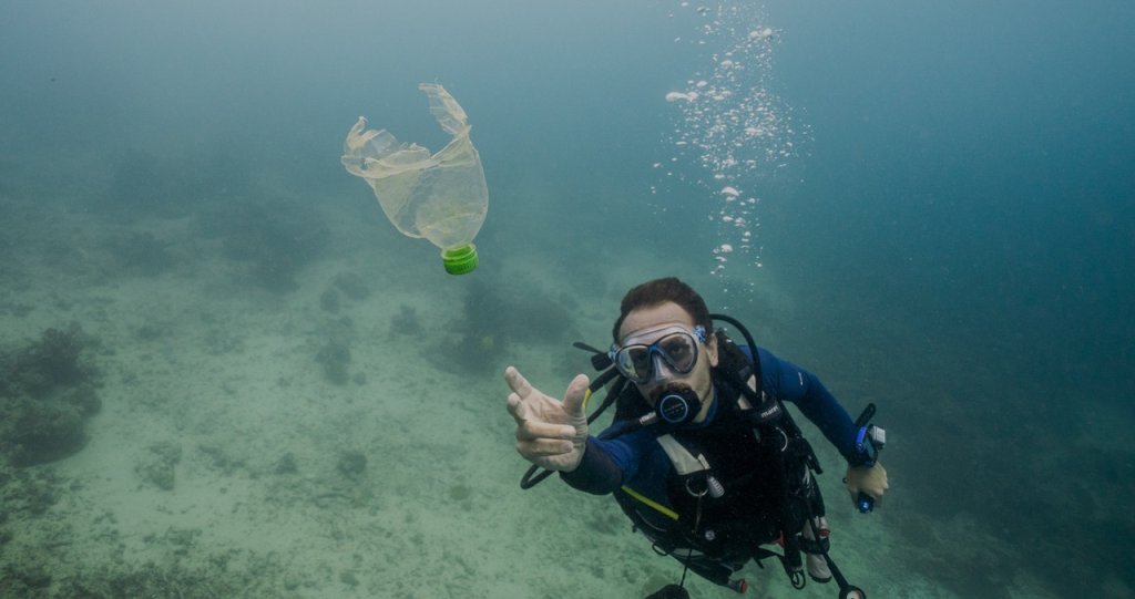 A scuba diver underwater reaches for a floating plastic bottle with marine life visible beneath.