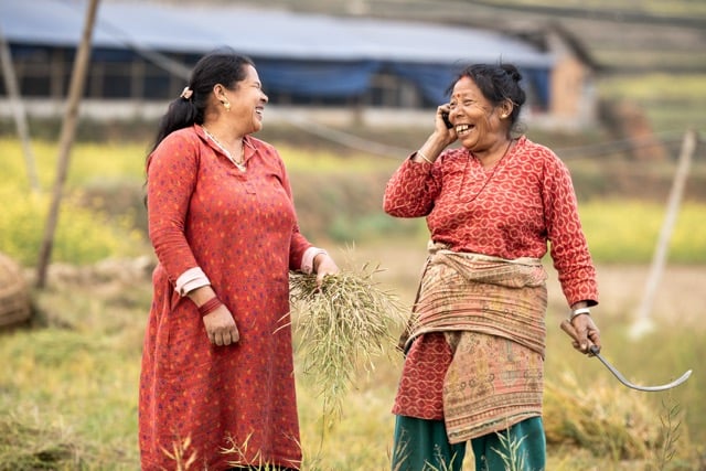 A woman in a red shirt and dark vest, representing GSMA Mobile for Development, is focused on her smartphone. Her long dark hair is tied back, accentuating her nose ring. The slightly blurred background reveals a textured wall and earthen ground, creating a soft, natural outdoor setting.