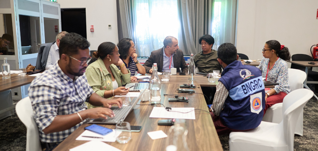 Seven people sit around a conference table in discussion, reviewing laptops, papers, and water bottles as they collaborate on early warning strategies.