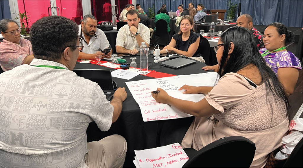 Seven people sit around a table discussing and writing ideas on large paper sheets during a group workshop.
