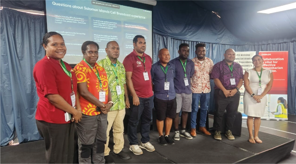 Nine people stand in a row onstage at an event, wearing lanyards, with a presentation screen behind them.