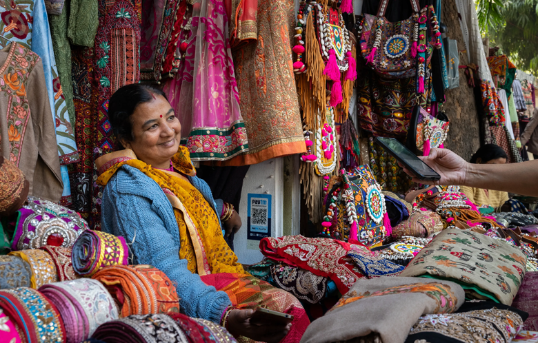 A woman smiles at a colourful market stall run by female micro-entrepreneurs, selling embroidered fabrics, bags, and garments.