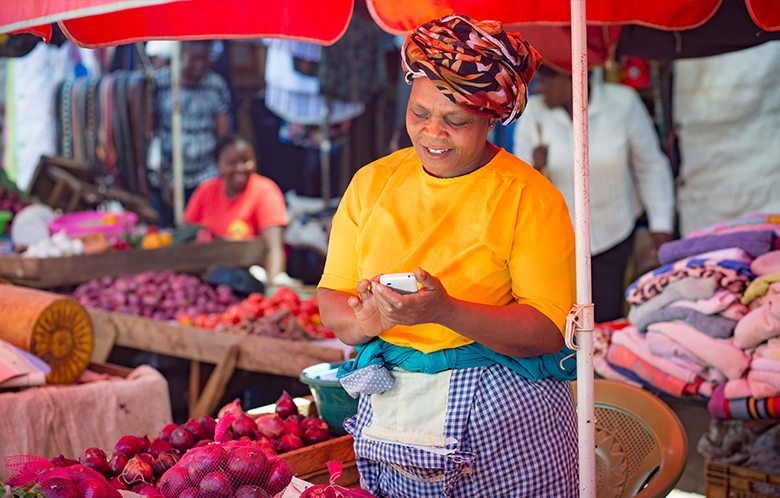 Smiling female micro-entrepreneur at a market stall uses her mobile, surrounded by onions and textiles under a red umbrella.