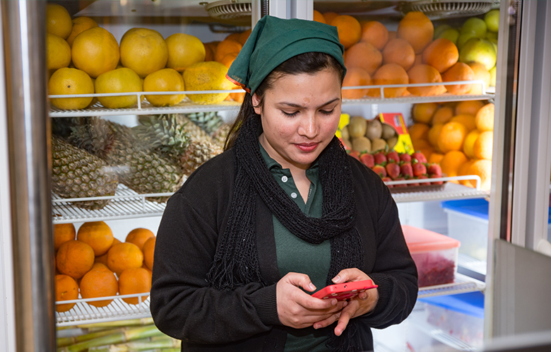 A female micro-entrepreneur stands in front of a fruit fridge, using her mobile as she's surrounded by oranges, pineapples, and grapefruits.