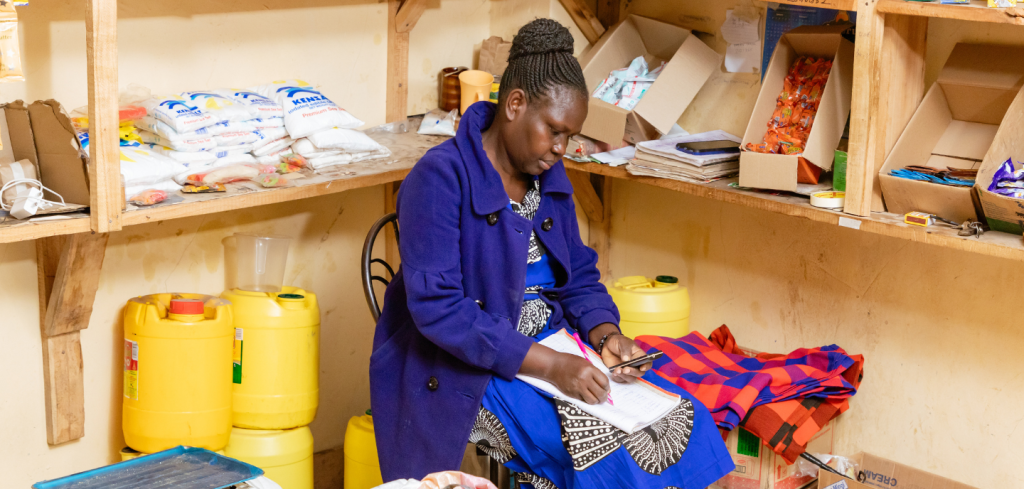 A woman in a blue coat sits on a chair, writing in a notebook on her lap. She is in a small shop with shelves holding food items, yellow jerrycans, checked cloth, boxes, and various packets against beige walls.