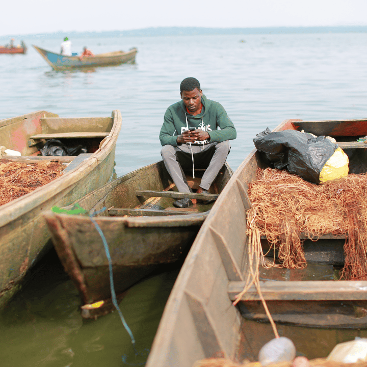 A man sits on the edge of a wooden boat on a calm lake, looking down at something in his hands. Fishing nets and bags are visible in the boat beside him. Other boats and people are seen in the background on the water.