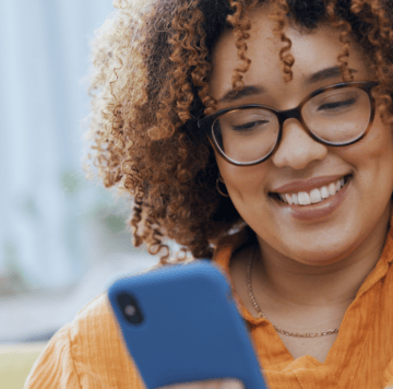 A woman with curly hair and glasses smiles whilst looking at her mobile. She is wearing a gold necklace and an orange top. The mobile has a blue case, and the background is softly blurred with natural light.