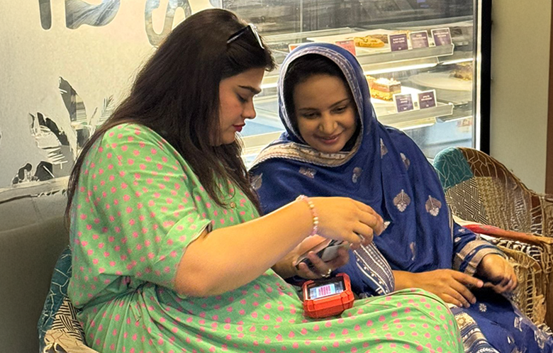 Two female micro-entrepreneurs sit together, smiling and looking at a mobile in a bakery or café setting.