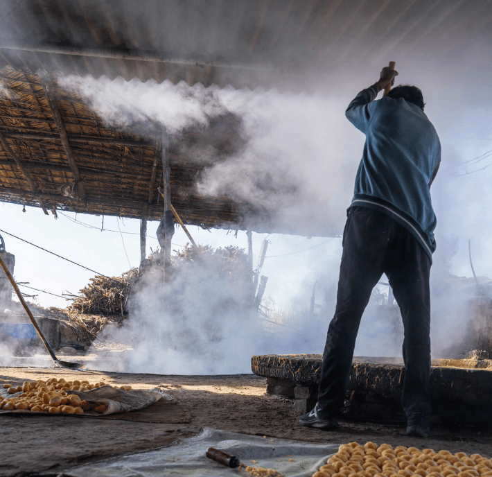 A person stands under a rustic shelter, raising an arm whilst surrounded by thick white smoke. Sunlight filters through the thatched roof, illuminating trays of yellow spiral snacks—an age-old process now merging with emerging ClimateTech solutions.