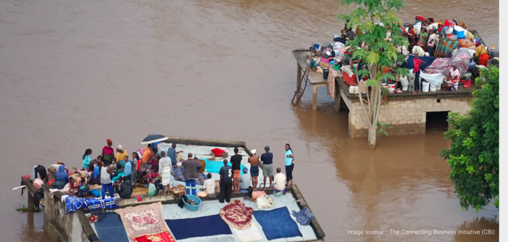 Dozens of people crowd on the roofs of two flooded buildings surrounded by muddy water, with belongings like blankets and bags. A tree is near one building. The floodwater covers the ground, leaving only rooftops accessible.