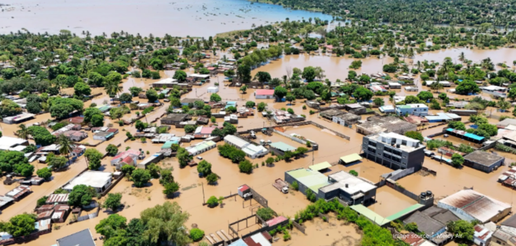 Aerial view of a flooded town with muddy water submerging streets and buildings. Numerous homes and trees are partially underwater, and large sections of the area are surrounded by floodwaters extending into distant fields.