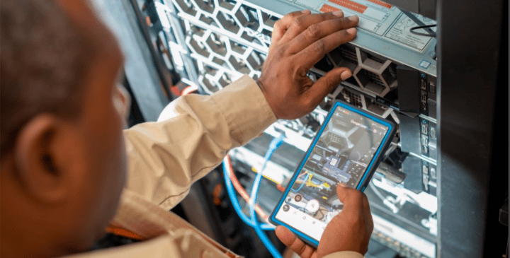 A person in a beige shirt uses one hand to adjust a server rack and the other to hold a mobile phone, showcasing Mobile for Humanitarian Innovation. The phone displays equipment details, with colourful cables visible in the data centre environment.