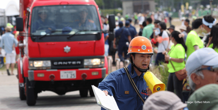 A worker in a blue uniform and orange helmet holds a yellow megaphone and a clipboard in front of a red fire engine, coordinating digital humanitarian assistance as a crowd in bright clothing gathers at an outdoor event.