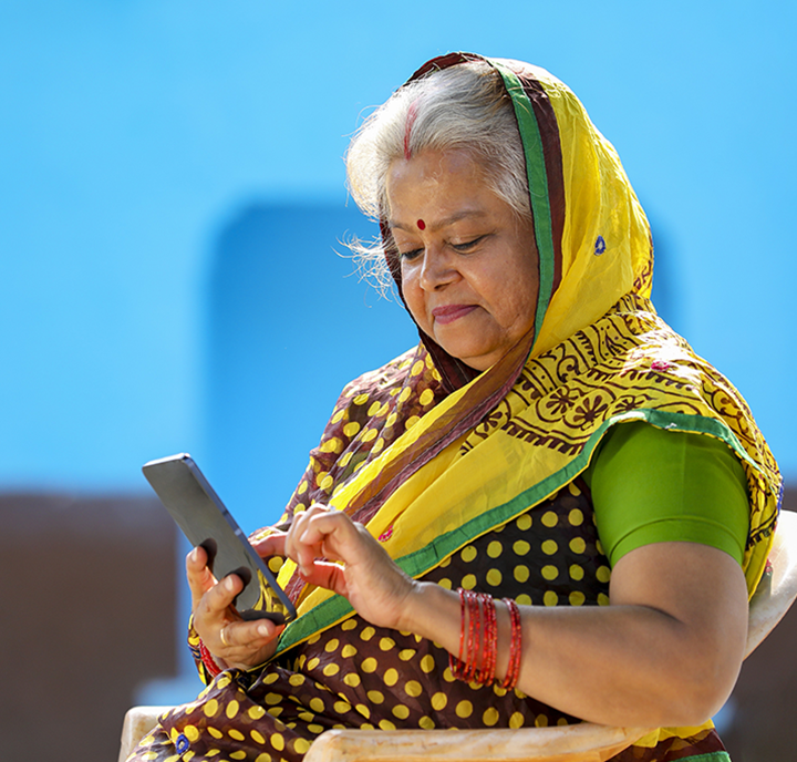 An elderly woman in a colourful sari sits outdoors, smiling as she uses a smartphone—a true symbol of Connected Women. She has grey hair, red bangles, and a red bindi; the bright blue background adds vibrancy to the scene.