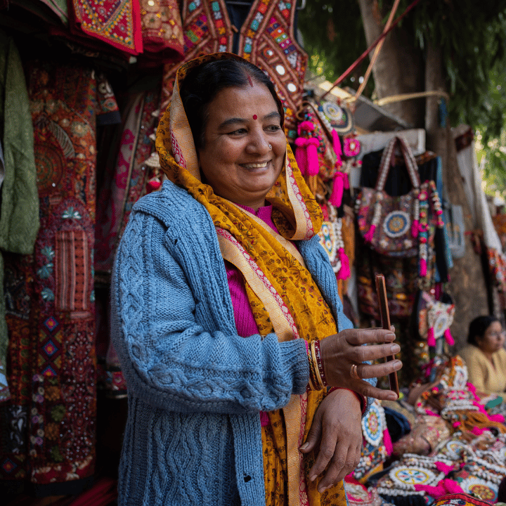 A smiling woman in a blue jumper and yellow dupatta stands at a vibrant outdoor market stall, surrounded by colourful, embroidered textiles, bags, and wall hangings. She holds a stick in one hand, with trees and another person visible in the background.