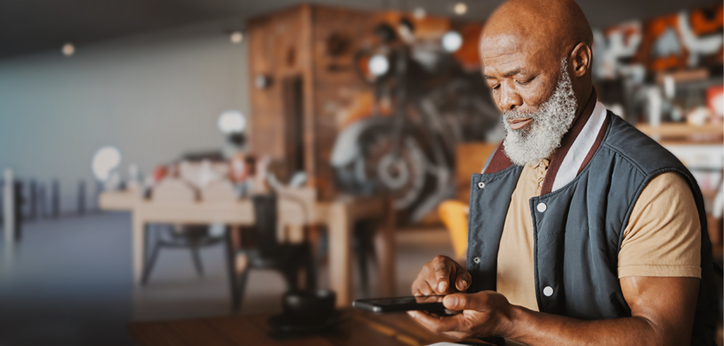 An older man with a grey beard sits at a table in a cosy café, using a tablet. He wears a light brown shirt and a dark waistcoat. The background shows blurred tables, chairs, and warm décor, suggesting a relaxed, inviting atmosphere.