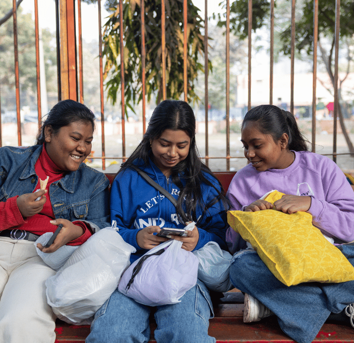 Three young women sit closely on a bench outdoors, smiling and looking at a mobile. They each hold bags or cushions and wear casual clothes: a denim jacket, a blue hoodie, and a lavender sweatshirt. Iron railings, trees, and blurred people are in the background.