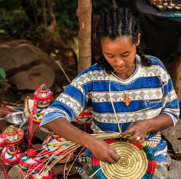 A woman with plaited hair weaves a colourful basket from natural fibres. She wears a blue and grey patterned shirt and a cross necklace. Finished woven baskets with red, green, and white designs are beside her. She works outdoors, surrounded by greenery and rocks.