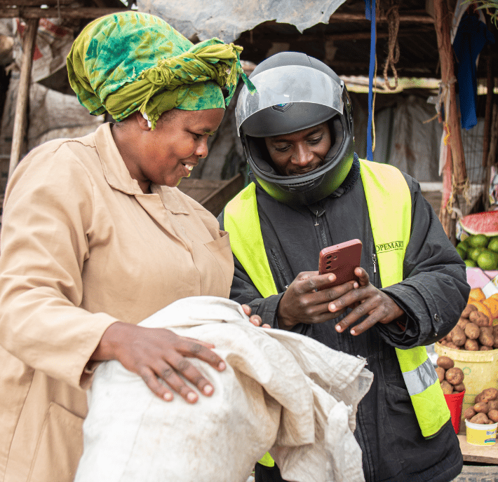 A smiling woman in a tan coat and green headscarf holds a large sack while a man in a black helmet and reflective vest, possibly working for local utilities, shows her something on his mobile at an outdoor market with fruit and vegetables in the background.