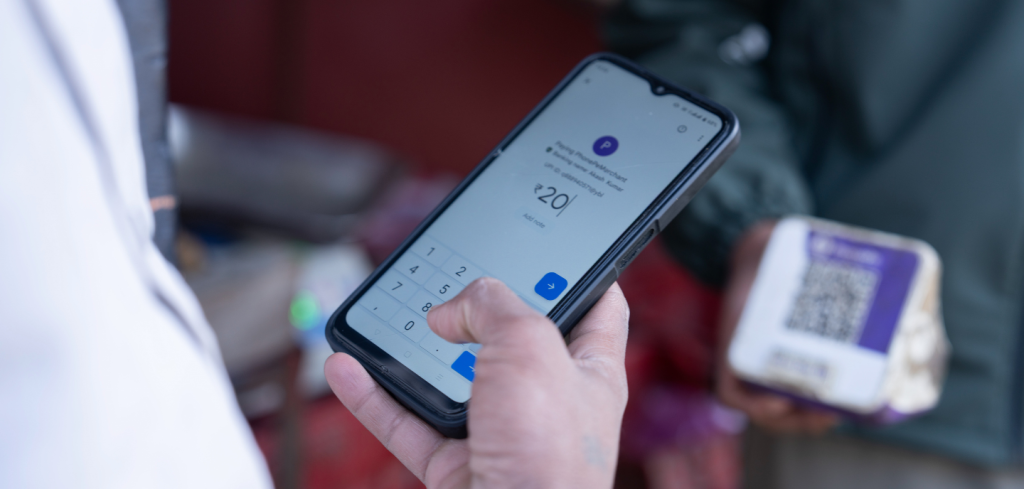 A person’s hand holds a mobile phone showing a digital payment app with ₹20 entered on the keypad. Another person holds a product and a box displaying a QR code, suggesting a cashless transaction at a market or shop.