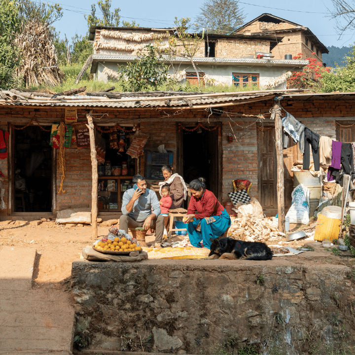 A family of four sits outside a rustic brick house, with sacks, goods, and laundry hanging nearby. A woman tends to items on a mat, a man eats, a child sits between them, and a black dog sleeps in front. Bright sunlight and greenery frame the rural scene.